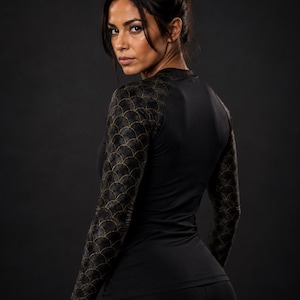 May include: A long-sleeve athletic shirt featuring a solid black torso and sleeves adorned with a gold-colored scale pattern. The garment has a crew neckline and a fitted silhouette, constructed from a smooth, dark fabric with metallic detailing.