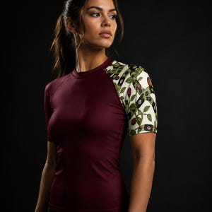 Op de afbeelding: Een sportshirt met korte mouwen, een effen diepbordeauxrode romp en raglanmouwen met een crèmekleurige botanische print. Het patroon toont groene bladeren, bruine stengels en kleine rode bessen in een herhalend geometrisch ontwerp.