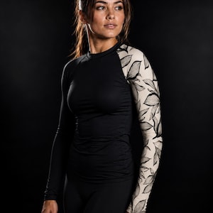 Op de afbeelding: Een sportshirt met lange mouwen, een effen zwarte romp en rechtermouw, gecombineerd met een linkermouw in beige met een zwart lijnpatroon van botanische bladeren. Het kledingstuk heeft een ronde halslijn en een nauwsluitende pasvorm.