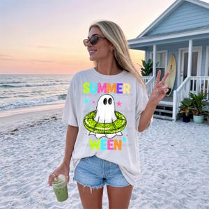 May include: A cream-coloured t-shirt with the words "SUMMER WEEN" in bright text. The shirt features a cartoon ghost wearing a green and yellow inflatable ring. The person is wearing blue denim shorts and sunglasses.