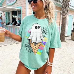 Puede incluir: Camiseta verde claro con un diseño de cono de helado con forma de fantasma. El fantasma tiene ojos negros y está cubierto por un cono de gofre. Las palabras "ICE CREAM" están escritas en letras coloridas. Hay telarañas negras a ambos lados del cono.
