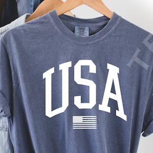 May include: A navy blue t-shirt with the word "USA" printed in white with a small American flag underneath. The shirt is made of a soft, comfortable material.