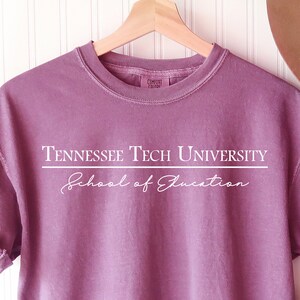 Pode incluir: Uma camiseta malva com mangas enroladas exibe o texto "Tennessee Tech University" acima de "School of Education" em branco. Um chap&eacute;u marrom e uma bolsa bege cheia de flores tamb&eacute;m s&atilde;o vis&iacute;veis. A camiseta est&aacute; pendurada em um cabide de madeira.