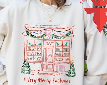 May include: Sand-colored sweatshirt featuring a festive illustration of a bookstore with the text "The Book Shop" and "A Very Merry Bookmas." The design includes Christmas lights, trees, and gift boxes, creating a holiday theme.