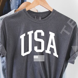 May include: A dark gray t-shirt with a white "USA" graphic and a small American flag underneath. The shirt is made of a soft, comfortable material.