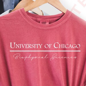 Pode incluir: Uma camiseta vermelha com o texto "UNIVERSITY OF CHICAGO" acima de uma linha horizontal e "Biophysical Sciences" abaixo. A camisa est&aacute; pendurada em um cabide de madeira com outras roupas e um chap&eacute;u marrom.