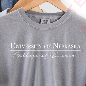 Pode incluir: Uma camiseta cinza com o texto "UNIVERSITY OF NEBRASKA College of Business" em branco. A camiseta est&aacute; pendurada em um cabide de madeira com uma camisa jeans e um chap&eacute;u marrom ao fundo.