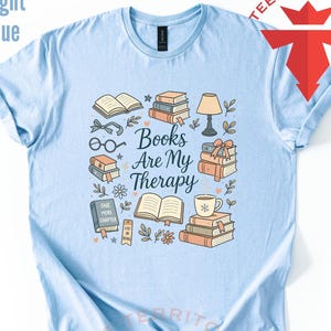 Może przedstawiać: Jasnoniebieska koszulka z graficznym wzorem przedstawiającym stosy książek, okulary, lampę i napis "Books Are My Therapy". Projekt zawiera elementy kwiatowe i gwiazd, tworząc przytulną estetykę związaną z książkami.