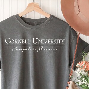 Pode incluir: Uma camiseta cinza com o texto "Cornell University Computer Science" em branco. A camisa est&aacute; pendurada em um cabide de madeira. Um chap&eacute;u marrom e uma bolsa bege cheia de flores tamb&eacute;m s&atilde;o vis&iacute;veis.