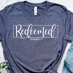 May include: A dark blue t-shirt with the word "Redeemed" in white script inside a white rectangle. The text "Ephesians 1:7" is printed below the rectangle.