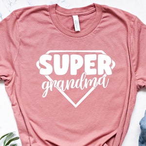 Może przedstawiać: Różowy t-shirt w kolorze pudrowego różu z białym nadrukiem w kształcie logo superbohatera i tekstem "SUPER grandma".