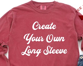 May include: A red long-sleeve t-shirt with the text "Create Your Own Long Sleeve" in white. The shirt is laid flat on a white surface with a pair of white sneakers and blue jeans.