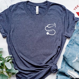 May include: A dark blue t-shirt with a white stethoscope and the word "nurse" printed on the front. The shirt is laid out on a white marble surface with a sprig of greenery and a pair of blue jeans.