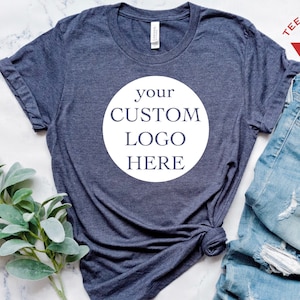 May include: A dark blue t-shirt with a white circle on the front that says "your custom logo here". The t-shirt is laid flat on a white marble surface with a sprig of greenery and a pair of blue jeans.