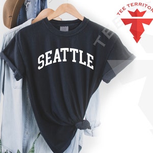 May include: Black t-shirt with white text that reads "SEATTLE" in a collegiate font. The shirt is hanging on a wooden hanger.