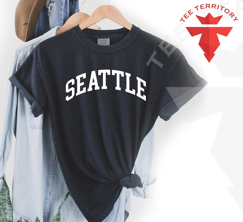 May include: Black t-shirt with white text that reads "SEATTLE" in a collegiate font. The shirt is hanging on a wooden hanger.
