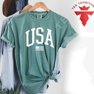 May include: A teal blue t-shirt with the word "USA" in white block letters and a small American flag graphic below.