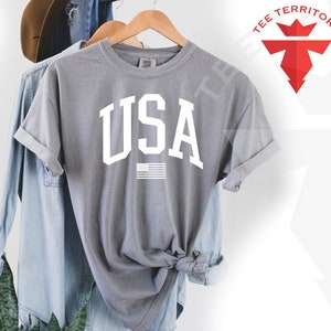 May include: A gray t-shirt with a white "USA" graphic and a small American flag underneath. The shirt is folded and displayed on a hanger.