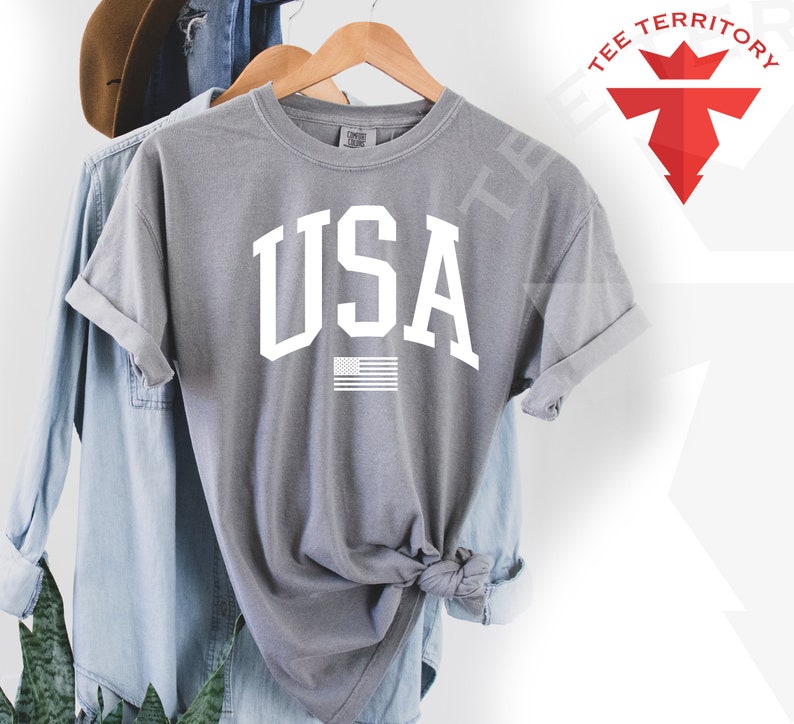 May include: A gray t-shirt with a white "USA" graphic and a small American flag underneath. The shirt is folded and displayed on a hanger.