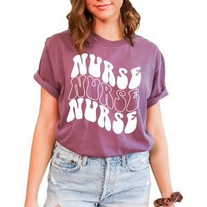 Puede incluir: Una camiseta morada con un gr&aacute;fico blanco que dice "NURSE" tres veces en una fuente retro y ondulada.