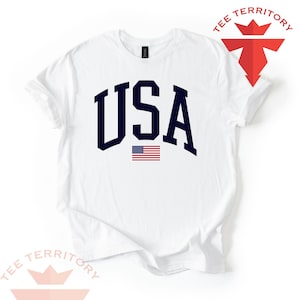 Puede incluir: Camiseta blanca con la palabra "USA" en letras may&uacute;sculas azul marino y una peque&ntilde;a bandera americana debajo. La camiseta tiene mangas cortas y cuello redondo. Las palabras "TEE TERRITORY" est&aacute;n impresas arriba a la derecha y abajo a la izquierda.