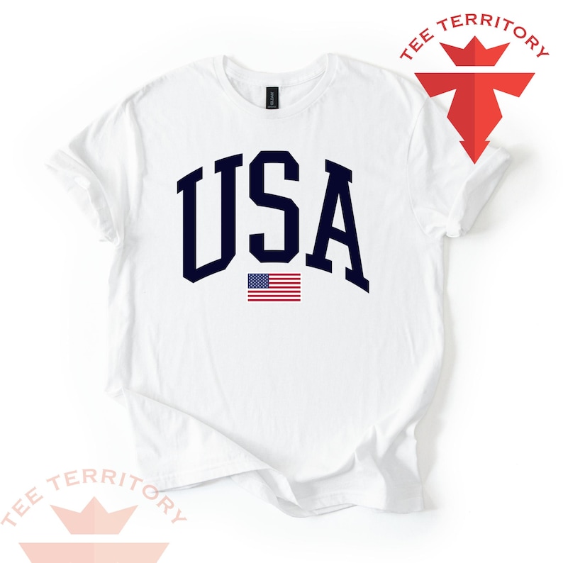 Puede incluir: Camiseta blanca con la palabra "USA" en letras may&uacute;sculas azul marino y una peque&ntilde;a bandera americana debajo. La camiseta tiene mangas cortas y cuello redondo. Las palabras "TEE TERRITORY" est&aacute;n impresas arriba a la derecha y abajo a la izquierda.