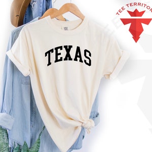 May include: A cream colored t-shirt with the word "TEXAS" printed in black on the front. The shirt is hanging on a wooden hanger.