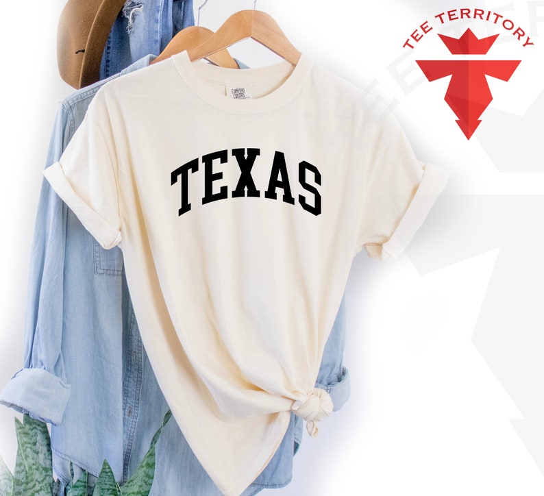 May include: A cream colored t-shirt with the word "TEXAS" printed in black on the front. The shirt is hanging on a wooden hanger.