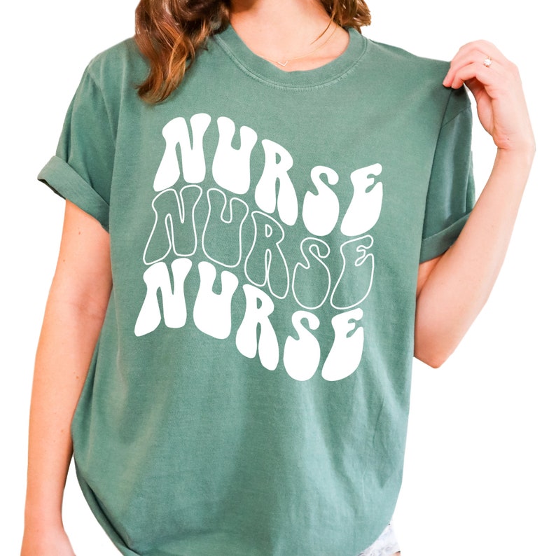 Puede incluir: Una camiseta verde azulada con un dise&ntilde;o gr&aacute;fico blanco que dice "NURSE" tres veces en una fuente retro y ondulada.