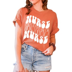 Puede incluir: Una camiseta de color terracota con un dise&ntilde;o gr&aacute;fico blanco que dice "NURSE" en una fuente retro y ondulada.