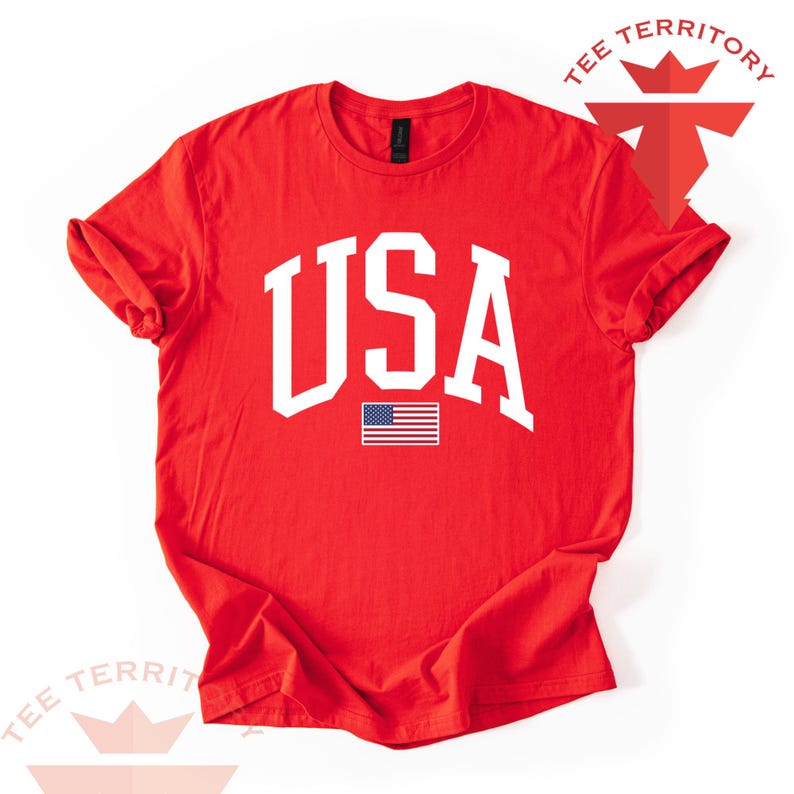 Puede incluir: Camiseta roja con la palabra "USA" en letras blancas grandes y una peque&ntilde;a bandera estadounidense. La camiseta tiene el texto "TEE TERRITORY" en las esquinas superior derecha e inferior izquierda.