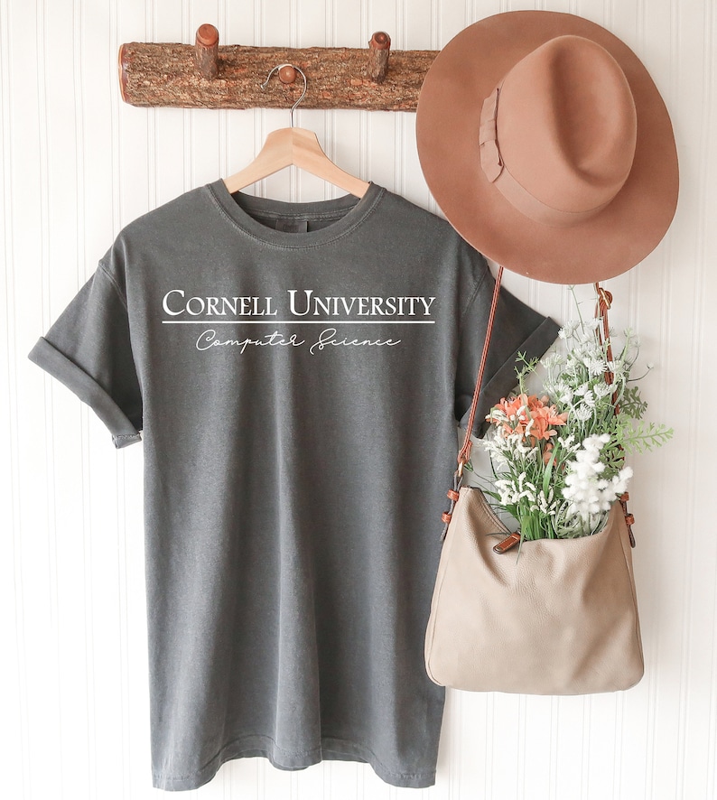 Pode incluir: Uma camiseta cinza com o texto "Cornell University Computer Science" em branco. A camisa est&aacute; pendurada em um cabide de madeira. Um chap&eacute;u marrom e uma bolsa bege cheia de flores tamb&eacute;m s&atilde;o vis&iacute;veis.