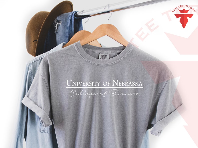 Pode incluir: Uma camiseta cinza com o texto "UNIVERSITY OF NEBRASKA College of Business" em branco. A camiseta est&aacute; pendurada em um cabide de madeira com uma camisa jeans e um chap&eacute;u marrom ao fundo.