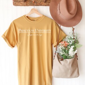 Pode incluir: Camiseta amarelo mostarda com o texto branco "PRINCETON UNIVERSITY Engineering". A camisa est&aacute; pendurada em um cabide de madeira. Um chap&eacute;u marrom e uma bolsa bege com flores tamb&eacute;m s&atilde;o vis&iacute;veis.
