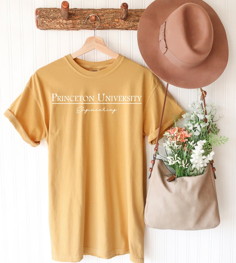 Pode incluir: Camiseta amarelo mostarda com o texto branco "PRINCETON UNIVERSITY Engineering". A camisa est&aacute; pendurada em um cabide de madeira. Um chap&eacute;u marrom e uma bolsa bege com flores tamb&eacute;m s&atilde;o vis&iacute;veis.