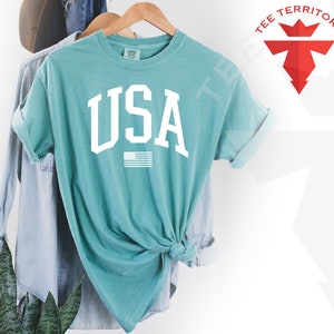 May include: A teal blue t-shirt with a white "USA" graphic and a small American flag underneath. The shirt is tied in a knot at the front.