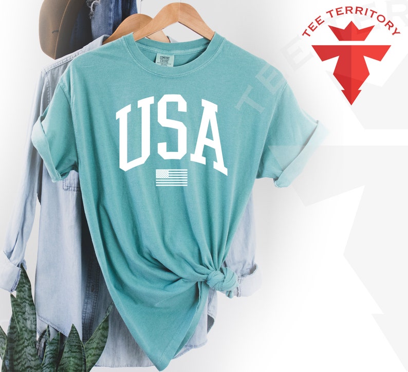 May include: A teal blue t-shirt with a white "USA" graphic and a small American flag underneath. The shirt is tied in a knot at the front.