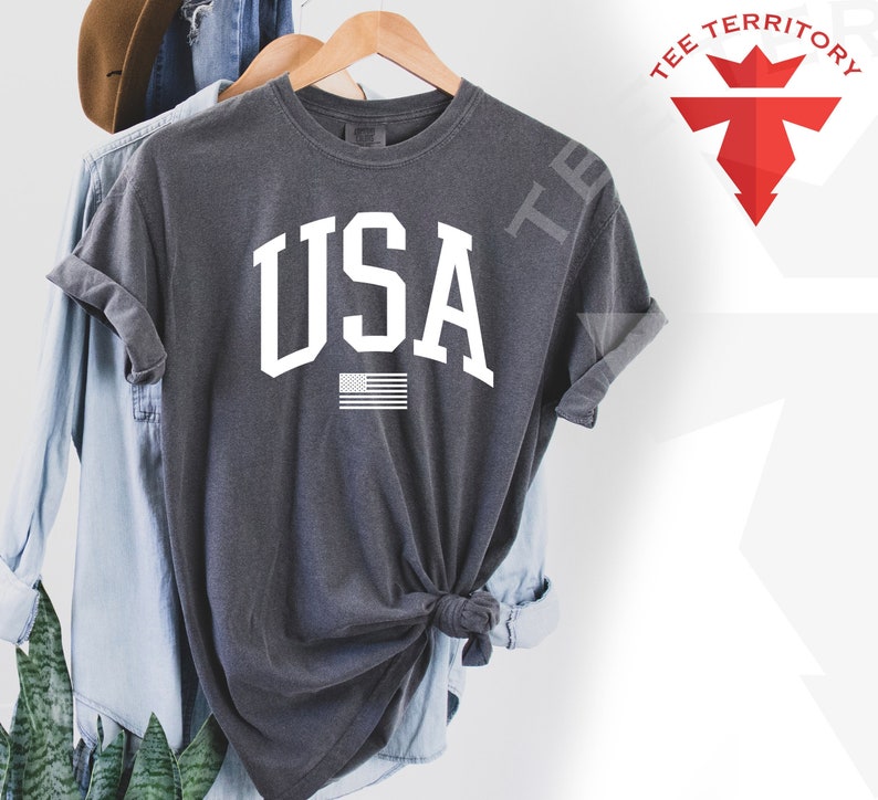May include: A dark gray t-shirt with a white "USA" graphic and a small American flag underneath. The shirt is made of a soft, comfortable material.