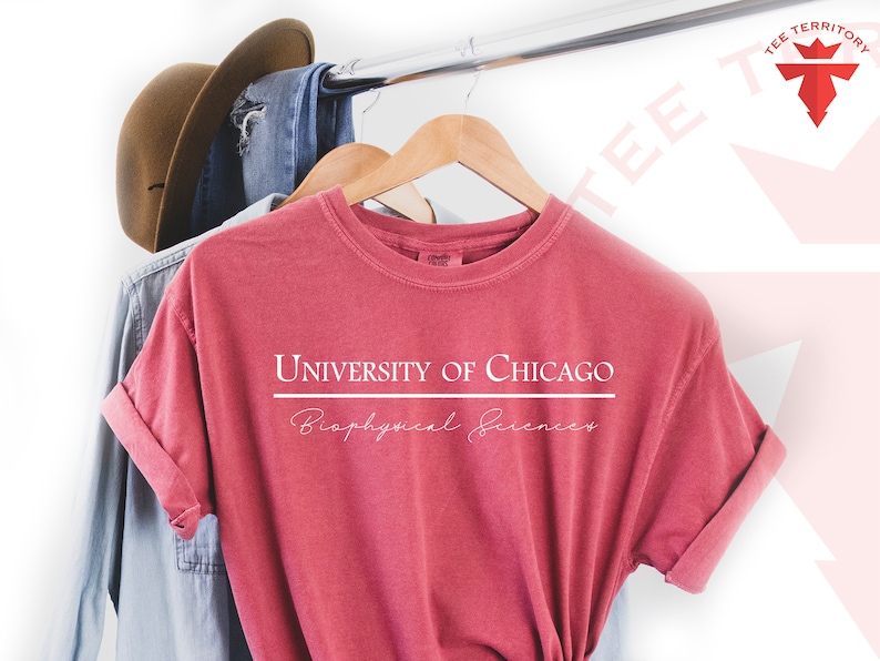Pode incluir: Uma camiseta vermelha com o texto "UNIVERSITY OF CHICAGO" acima de uma linha horizontal e "Biophysical Sciences" abaixo. A camisa est&aacute; pendurada em um cabide de madeira com outras roupas e um chap&eacute;u marrom.