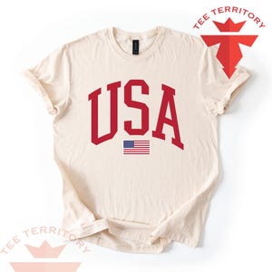 Puede incluir: Camiseta de color crema con la palabra "USA" en letras rojas y audaces de estilo universitario. Debajo del texto hay una peque&ntilde;a bandera estadounidense. La camiseta tiene cuello redondo y mangas cortas. El logotipo de la marca "TEE TERRITORY" es visible en la esquina superior derecha.