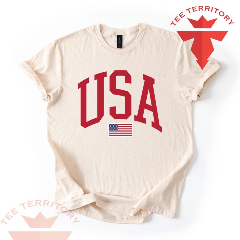 Puede incluir: Camiseta de color crema con la palabra "USA" en letras rojas y audaces de estilo universitario. Debajo del texto hay una peque&ntilde;a bandera estadounidense. La camiseta tiene cuello redondo y mangas cortas. El logotipo de la marca "TEE TERRITORY" es visible en la esquina superior derecha.
