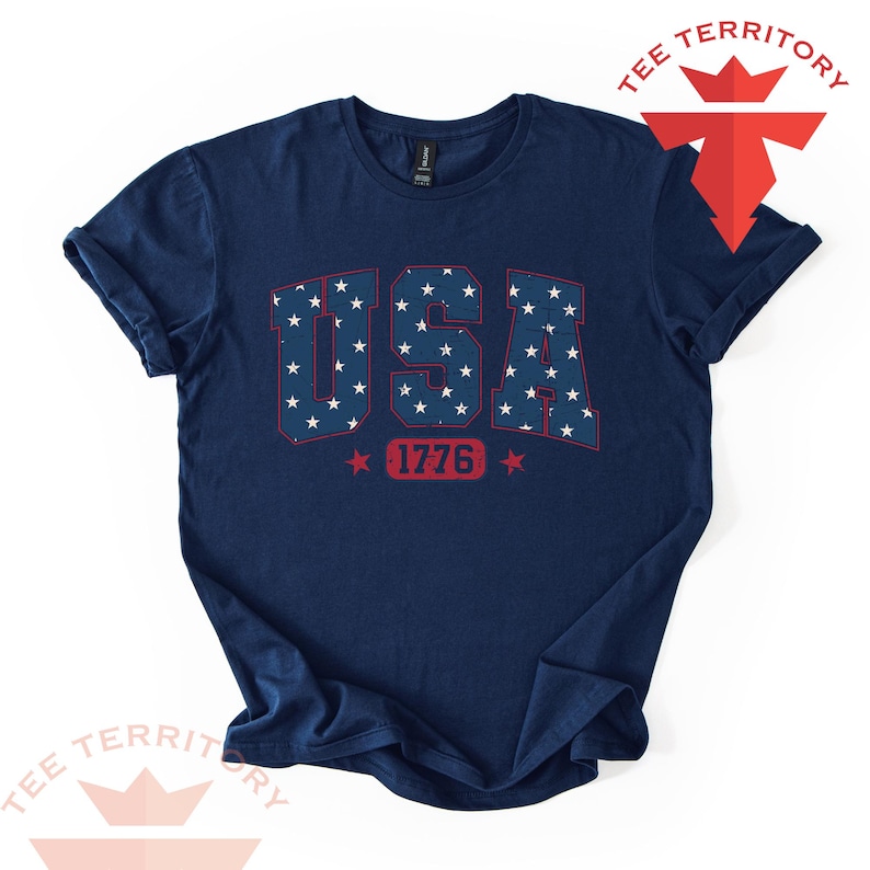 May include: Navy blue t-shirt with the word "USA" in red block letters filled with white stars. Below the word "USA" is the number "1776" in red. The shirt has rolled-up sleeves and a crew neck.