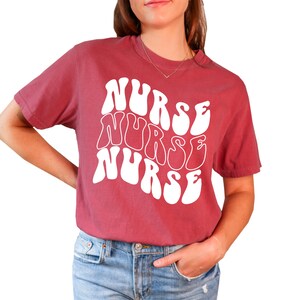Puede incluir: Una camiseta de color granate con un gr&aacute;fico blanco de la palabra "NURSE" repetida tres veces en una fuente ondulada y psicod&eacute;lica.