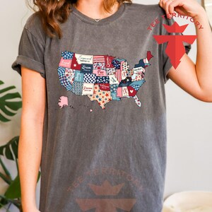 Pu&ograve; includere: Una maglietta grigia con un design a patchwork degli Stati Uniti. Ogni stato &egrave; un motivo diverso con parole come "Love", "Strong", "Grace", "Free", "Believe" e "Hope".