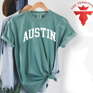May include: A teal blue t-shirt with the word "AUSTIN" printed in white on the front. The shirt is made of a soft, comfortable material and is perfect for everyday wear.