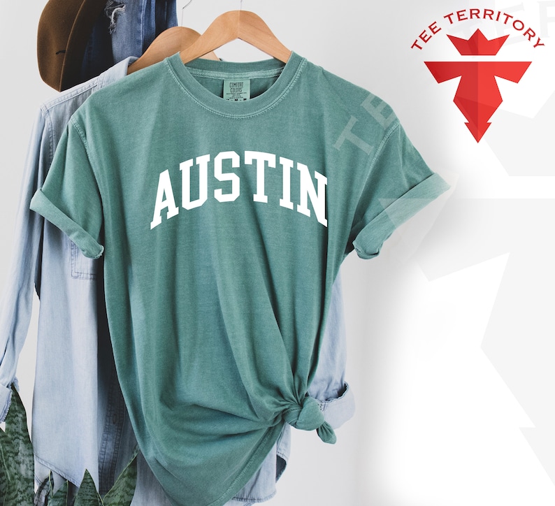 May include: A teal blue t-shirt with the word "AUSTIN" printed in white on the front. The shirt is made of a soft, comfortable material and is perfect for everyday wear.
