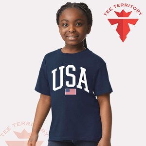 Puede incluir: Una camiseta azul marino con la palabra "USA" en blanco y una peque&ntilde;a bandera americana. La camiseta la lleva una persona joven. El logotipo de la marca "TEE TERRITORY" es visible.