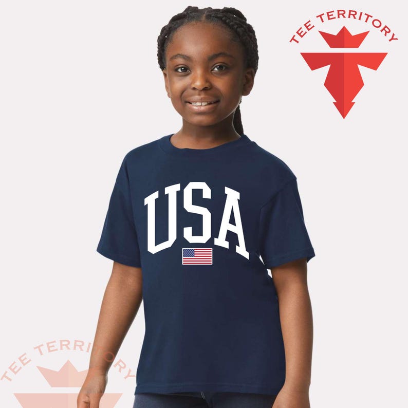 Puede incluir: Una camiseta azul marino con la palabra "USA" en blanco y una peque&ntilde;a bandera americana. La camiseta la lleva una persona joven. El logotipo de la marca "TEE TERRITORY" es visible.