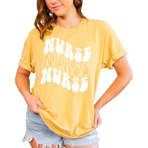 Puede incluir: Una camiseta amarilla con la palabra "NURSE" escrita tres veces en una fuente retro, blanca y ondulada.