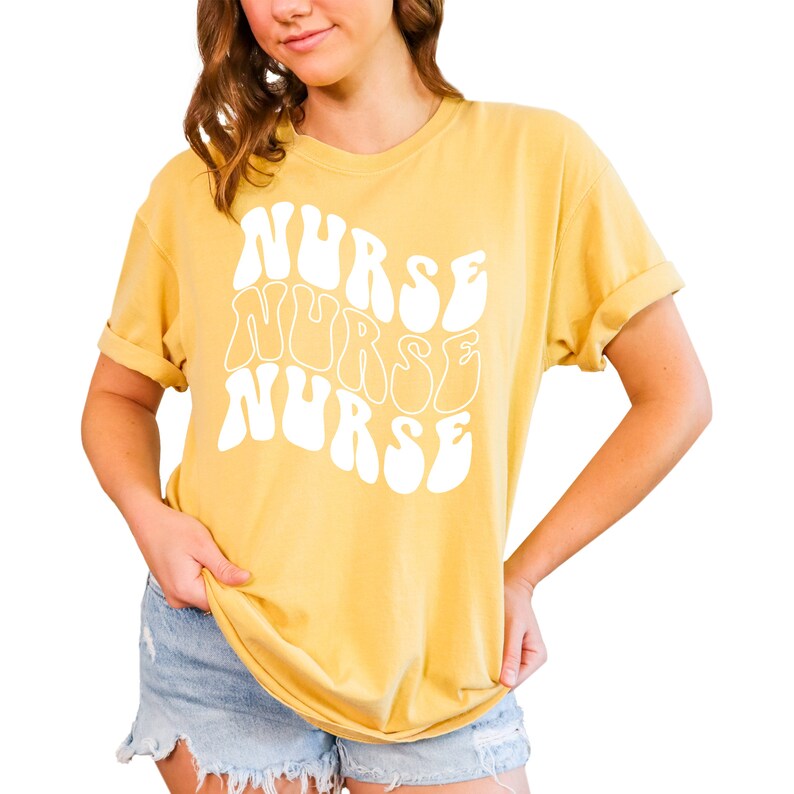 Puede incluir: Una camiseta amarilla con la palabra "NURSE" escrita tres veces en una fuente retro, blanca y ondulada.